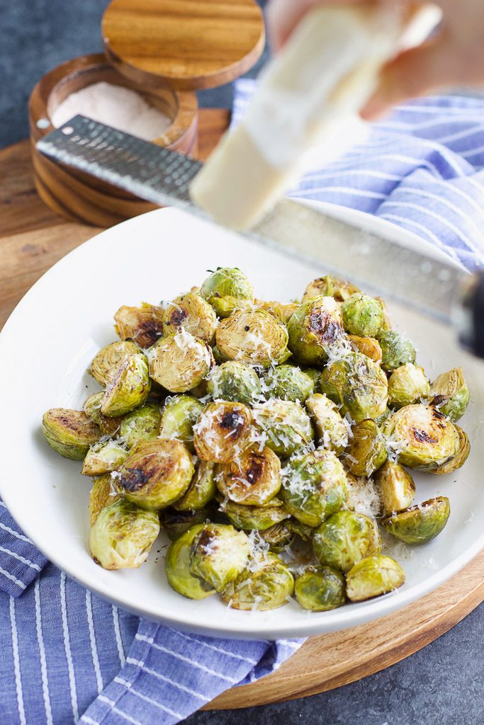 A hand grating cheese over a white bowl filled with brussels sprouts. 