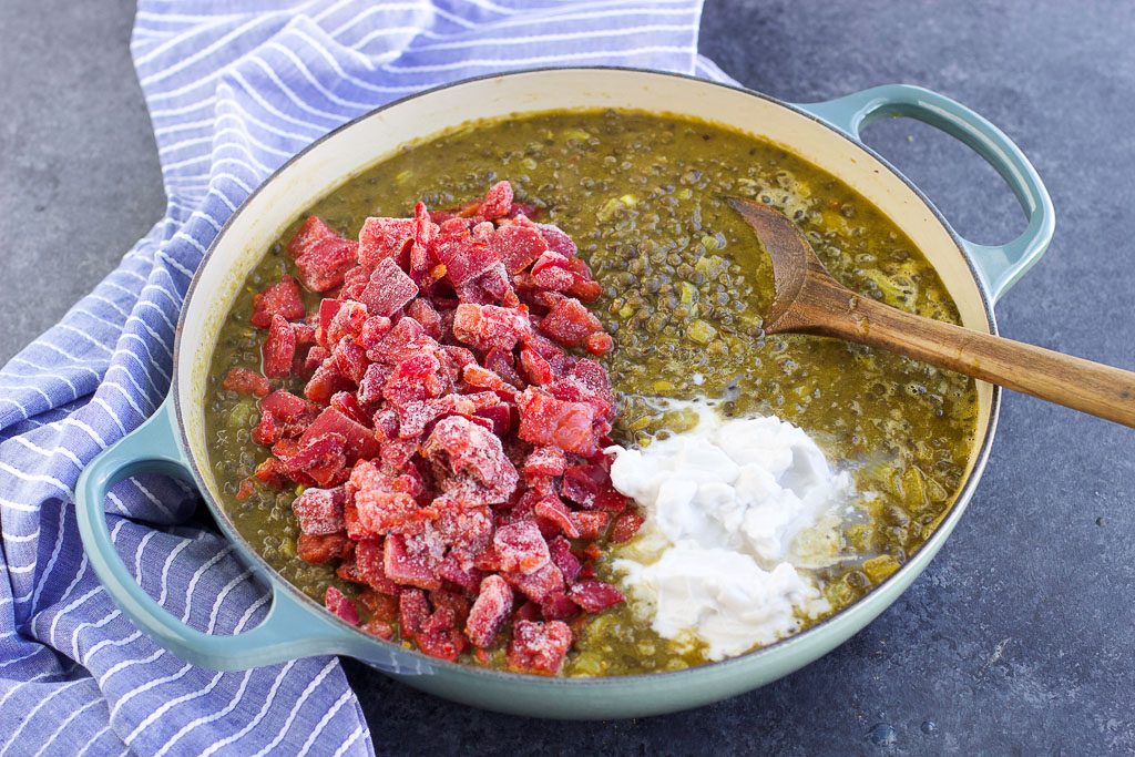 A large pan filled with lentils, curry paste, frozen red peppers, and coconut milk being stirred together with a wooden spoon.