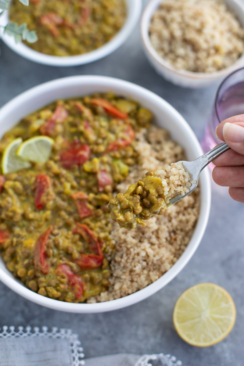 A hand holding a forkful of lentils over a white bowl on a gray background.