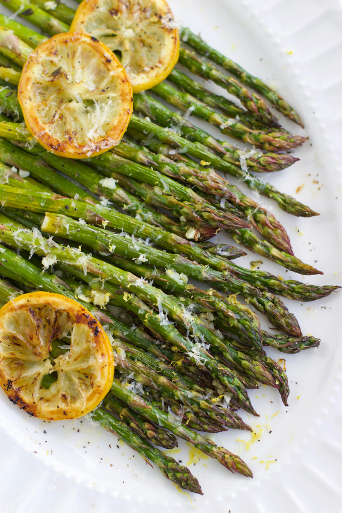 A large white platter filled with roasted asparagus topped with lemon slices and parmesan cheese. 