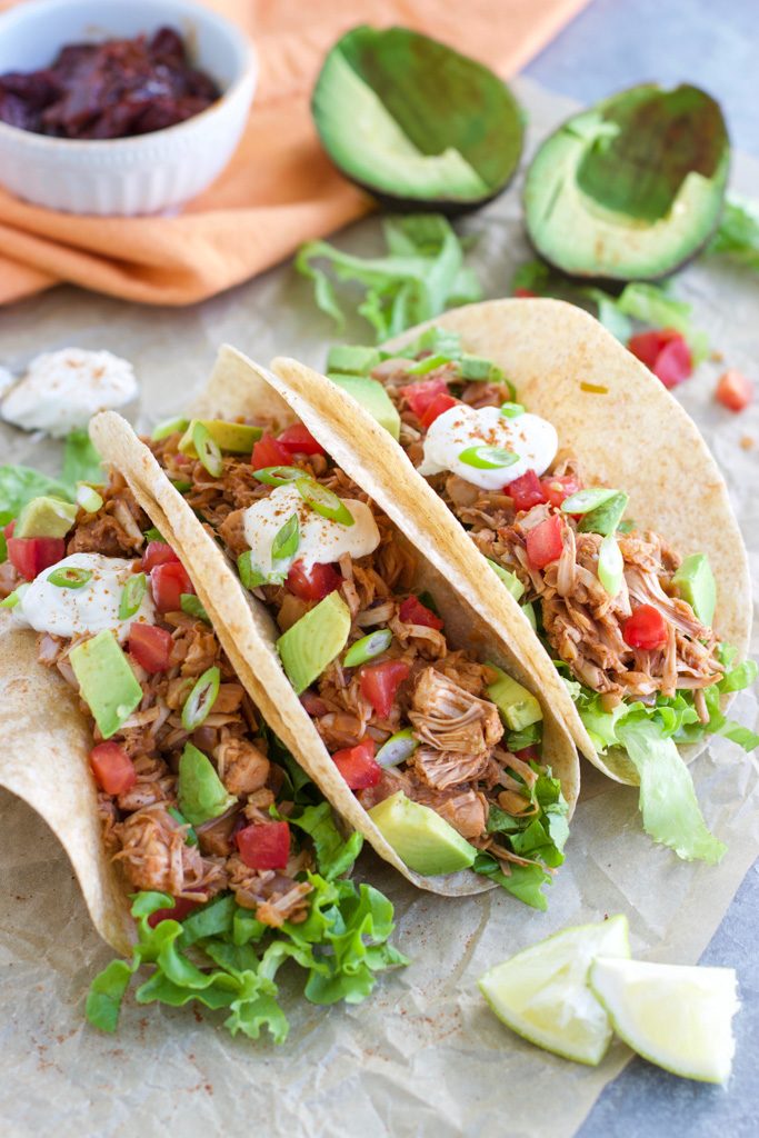 Three tacos resting on parchment paper surrounded by avocados, limes, and chipotle peppers.
