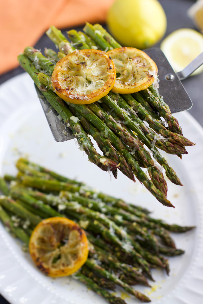 A metal spatula holding roasted asparagus with lemon slices over a large white platter. 