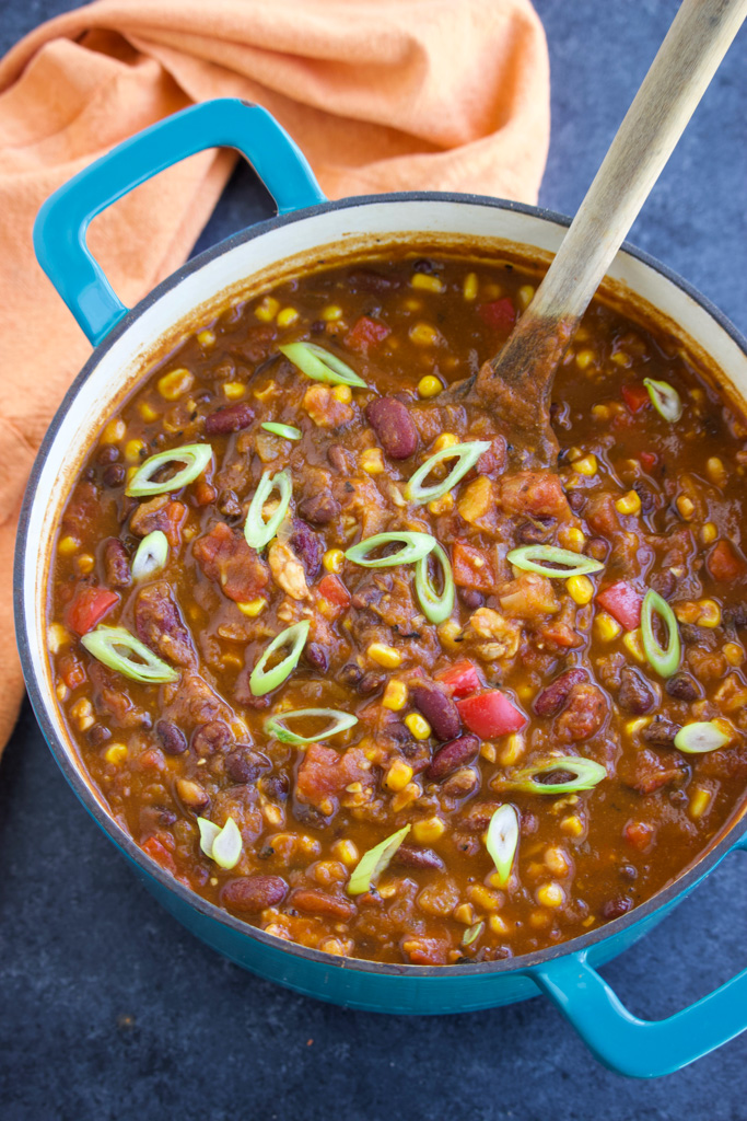 A blue pot filled with vegan chili and a wooden spoon next to an orange napkin on a dark background. 