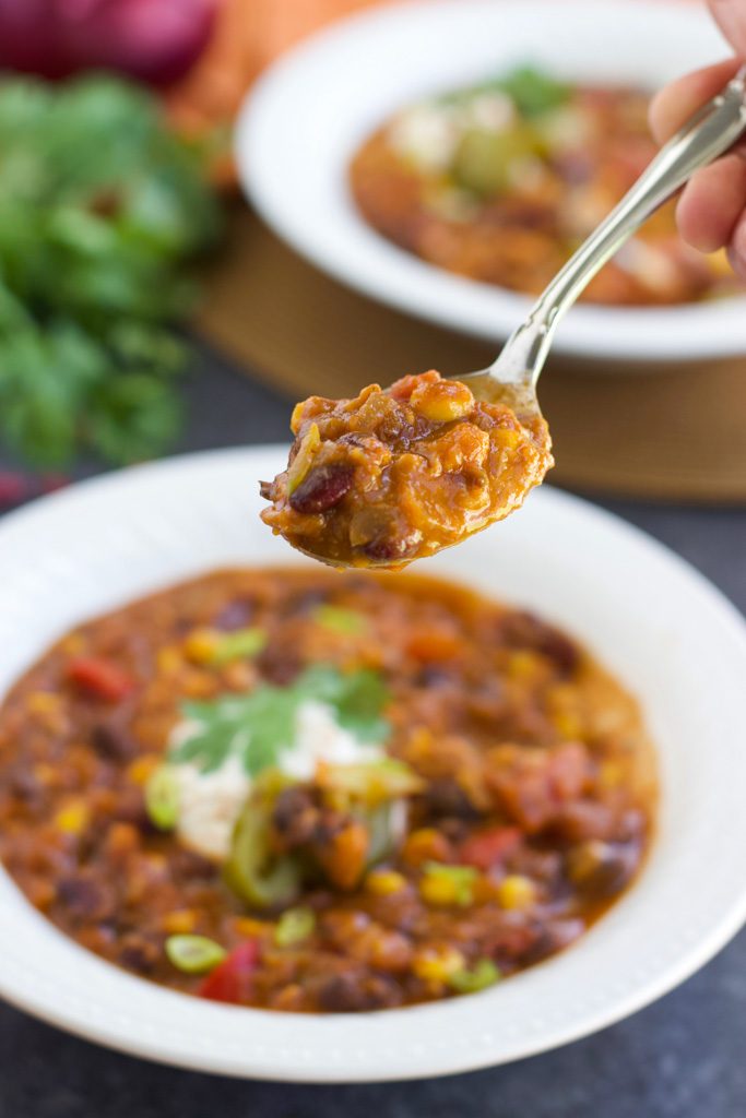 A hand holding a spoonful of chili over a white bowl with another bowl in the background.