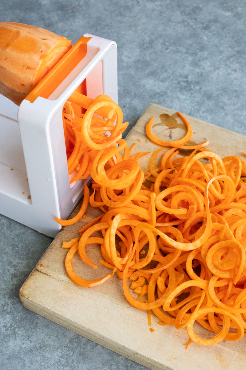 A sweet potato being spiralized into noodles onto a wooden cutting board on a gray background.