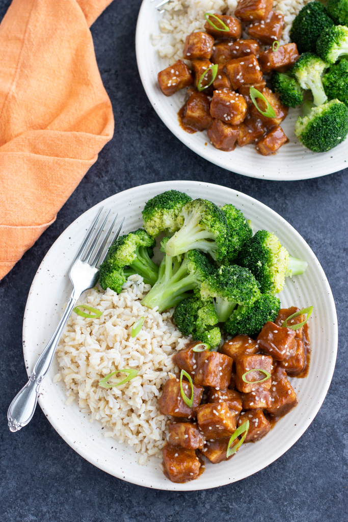 Two plates with tofu, broccoli, and brown rice on a dark background. 