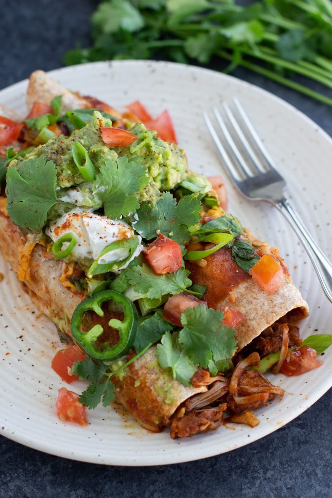 Two enchiladas topped with sliced jalapeños, guacamole, tomatoes, and cilantro on a white textured plate with a fork.