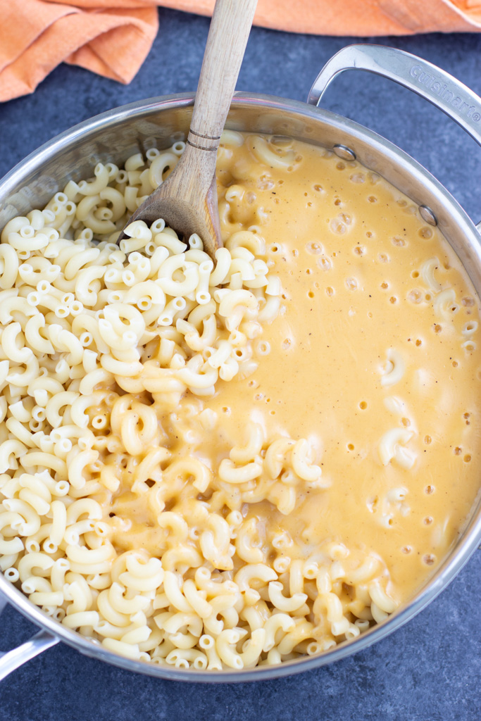 A wooden spoon stirring melted cheese with elbow noodles in a large pot on a dark background.