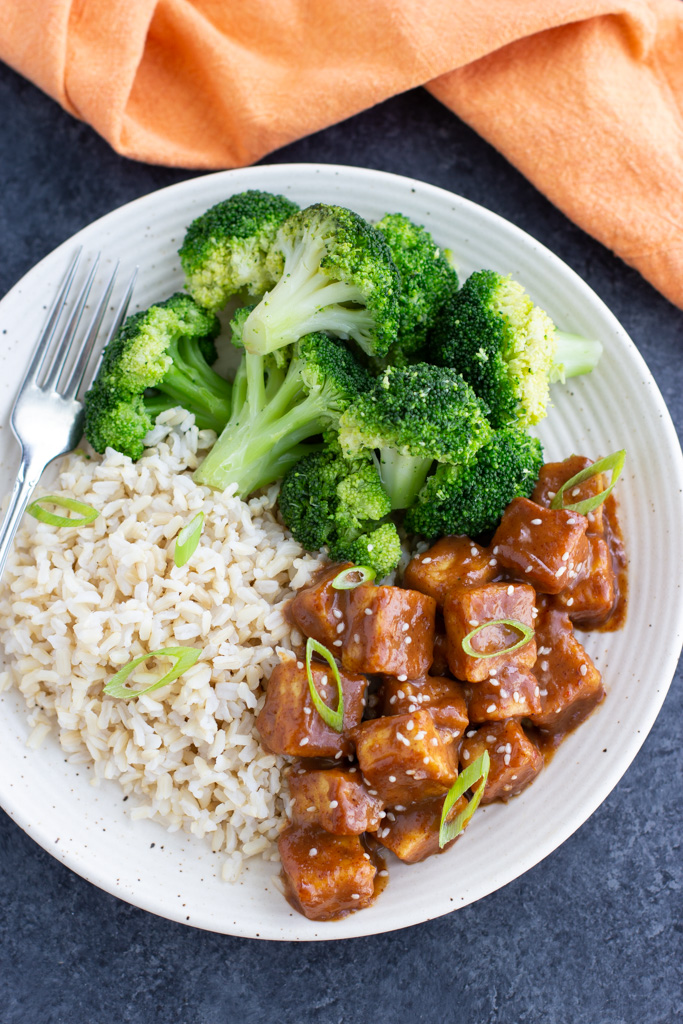 A white plate filled with tofu, brown rice, and broccoli florets next to an orange napkin on a dark background. 