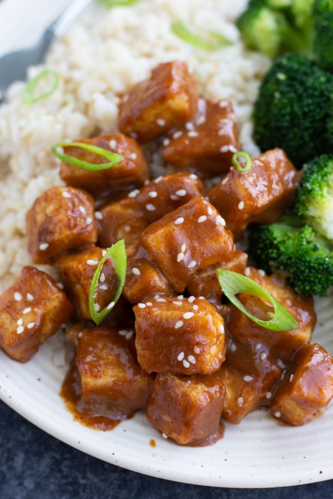 A close up view of a pile of tofu coated in thick almond butter sauce next to broccoli and rice on a plate.