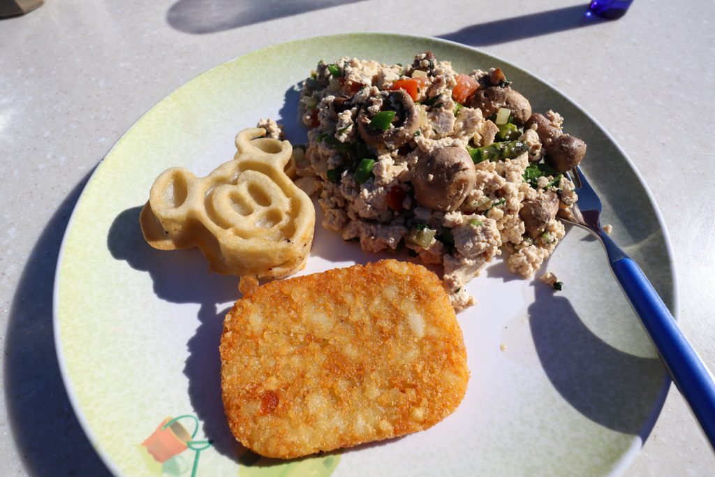 A plate with a Mickey waffle, tofu scramble, hash brown, and a fork on a table. 