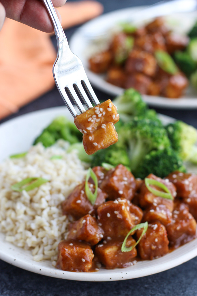 A hand holding a fork that's spearing two pieces of tofu over a plate of broccoli and rice.