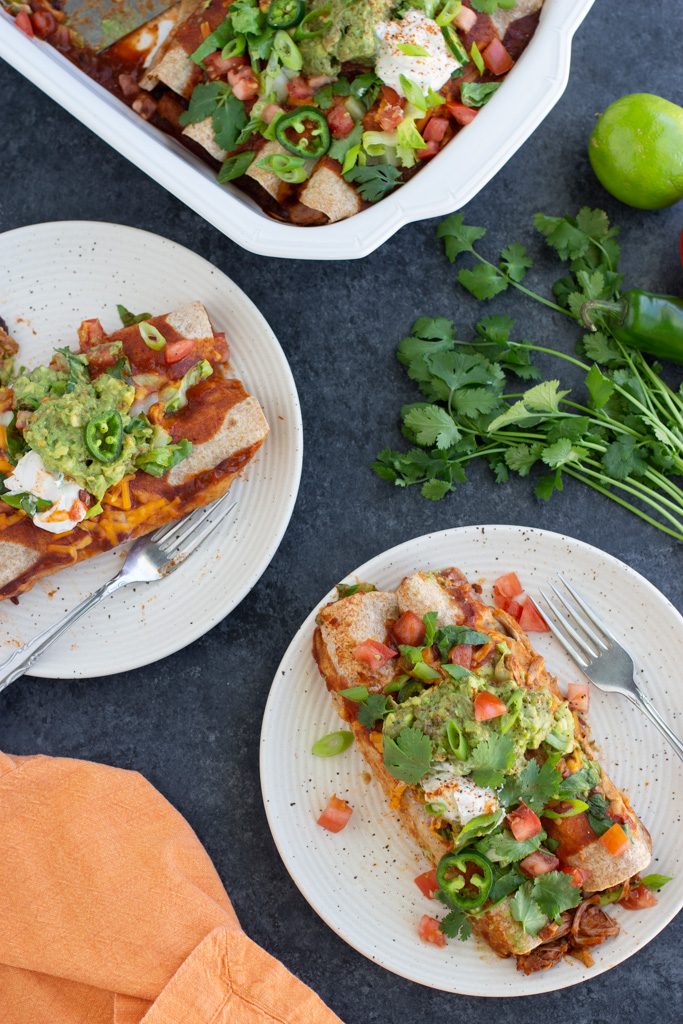 Overhead shot of enchiladas on two white plates next to a white casserole dish and fresh cilantro.