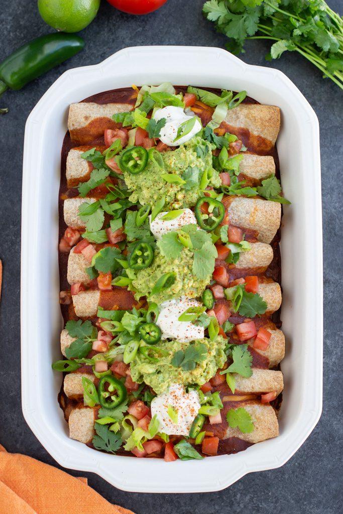 A large white casserole dish filled with jackfruit enchiladas and lots of toppings on a dark textured background.