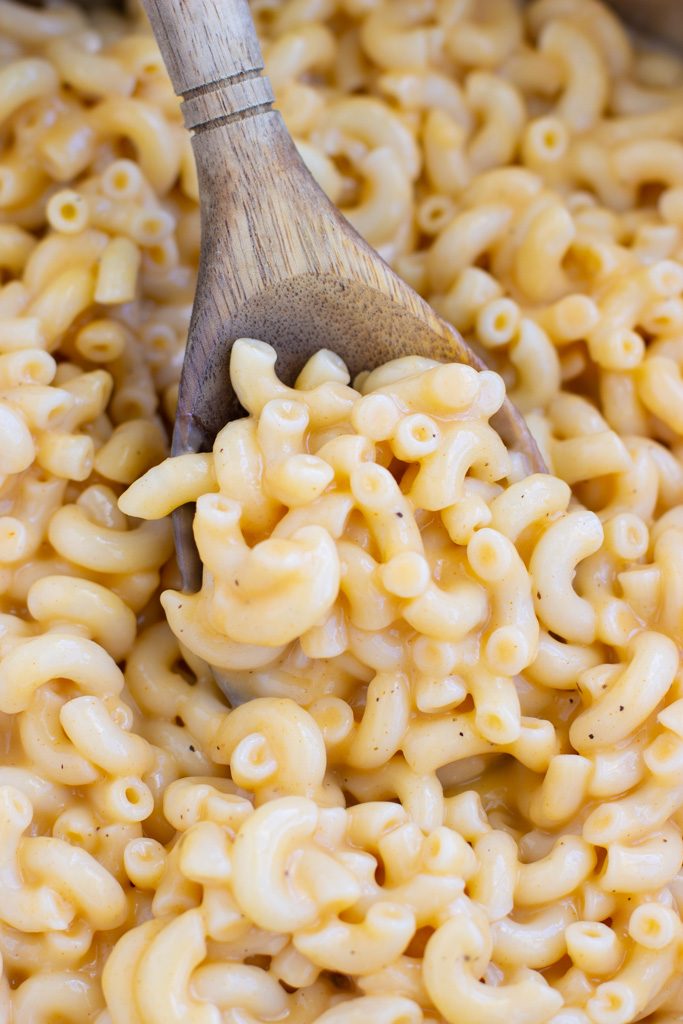Close up view of a wooden spoon scooping a large spoonful of cheesy noodles out of a pot. 