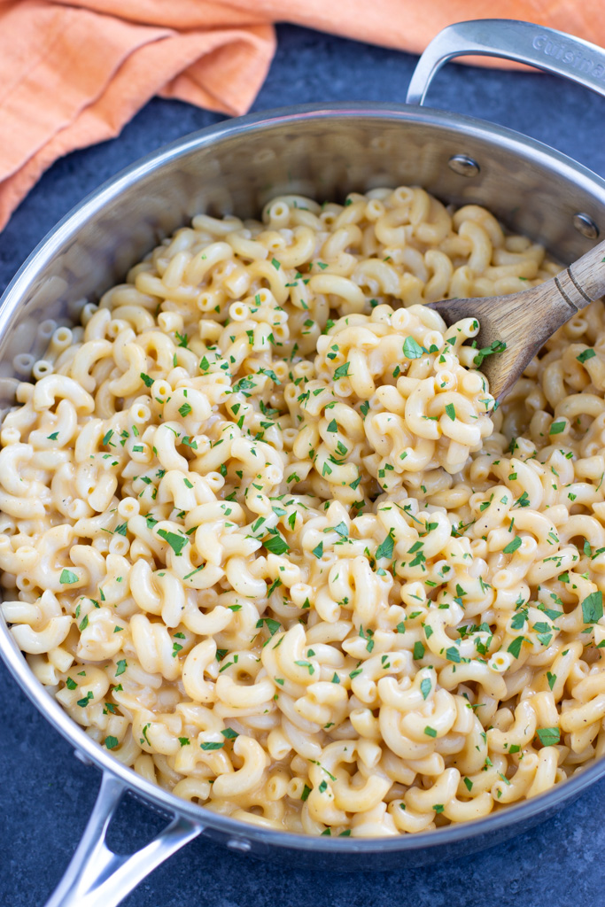 A large skillet of pasta garnished with parsley with a wooden spoon stirring it on a dark background. 