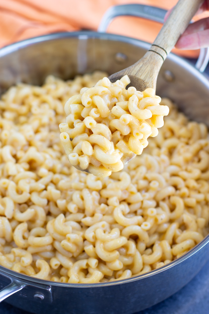 A wooden spoon scooping out a spoonful of macaroni and cheese out of a large silver pan next to an orange napkin. 