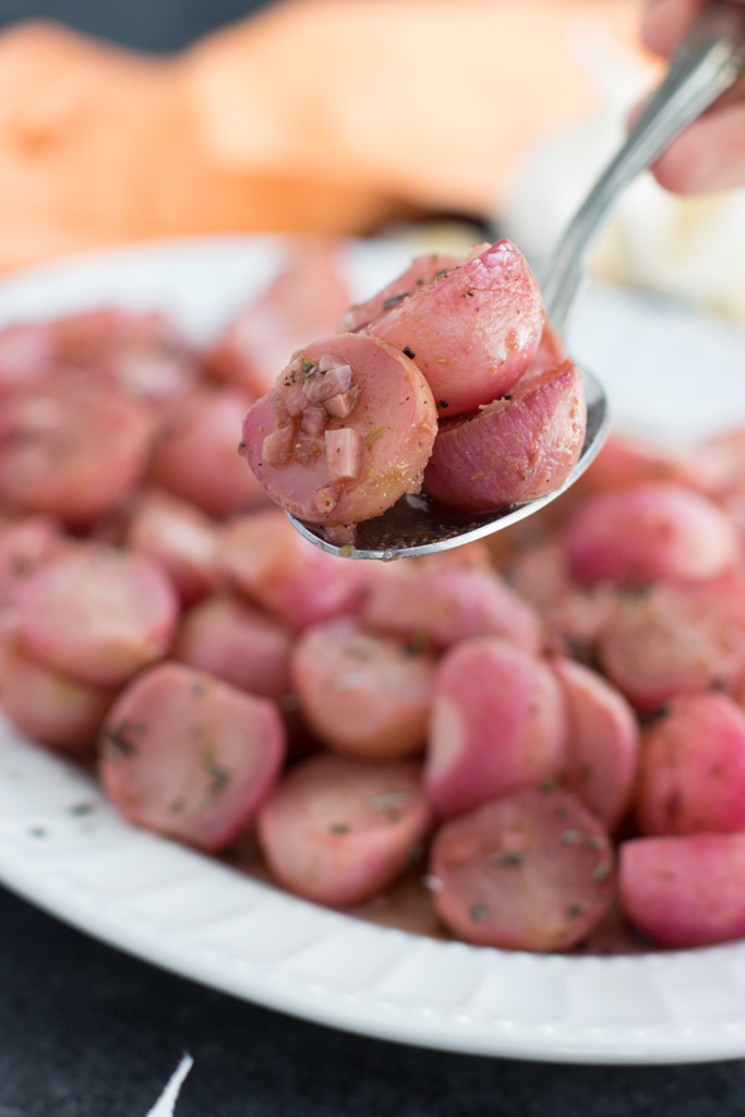 A hand holding up a large serving spoon of radishes over a white platter.