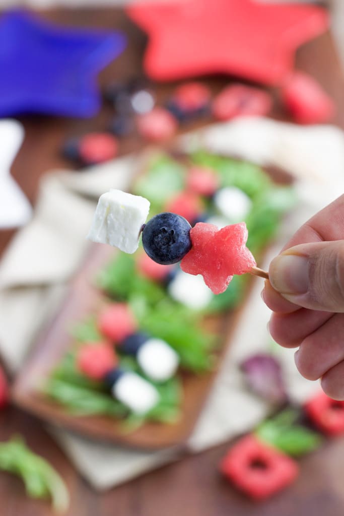 A hand holding a small red, white, and blue skewer in front of a platter of skewers.