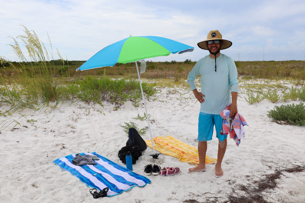 A man standing next to a beach umbrella with towels laid on the ground at Caladesi Island. 