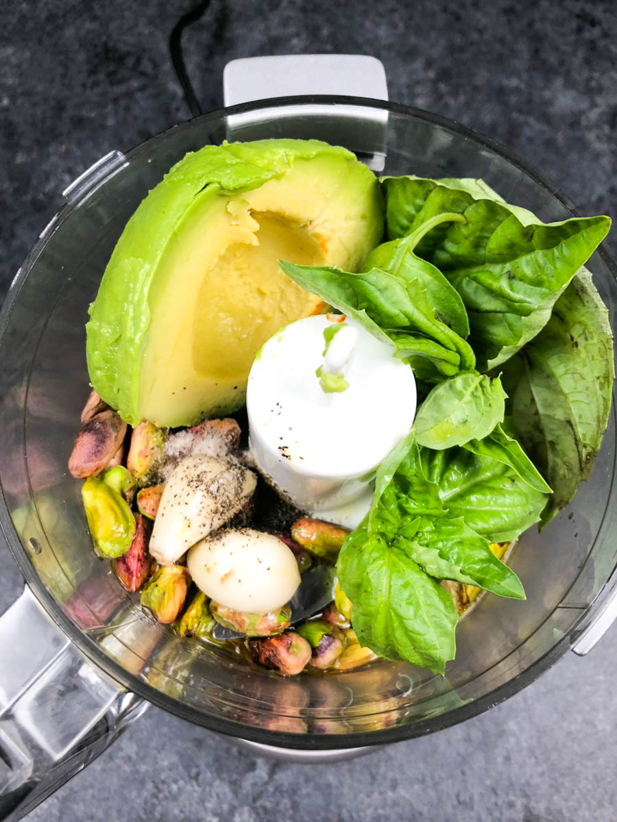 A food processor cup filled with avocado, basil, pistachios, and garlic on a dark background.