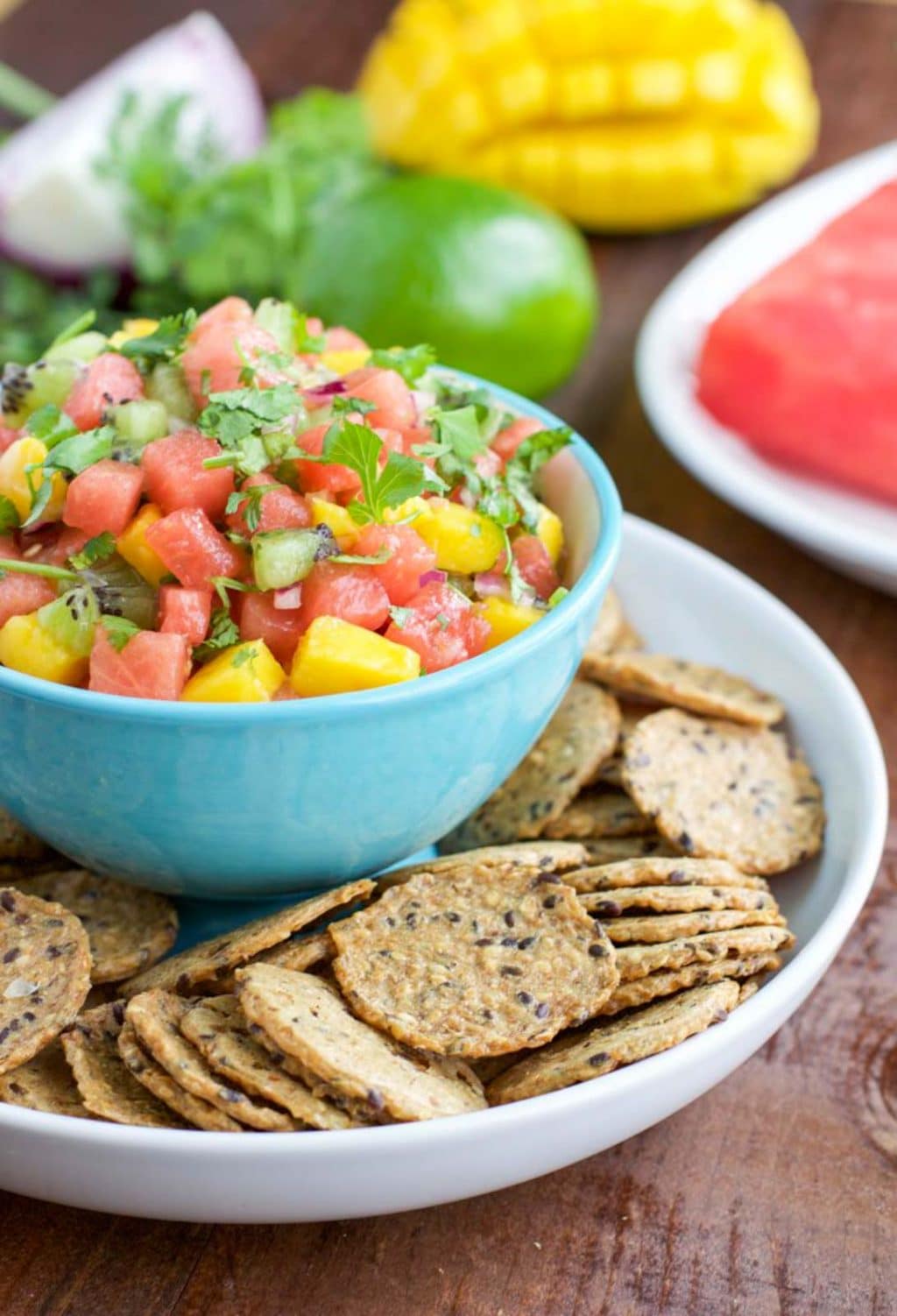 A large white platter filled with crackers surrounding a small bowl in the center that's filled with watermelon mango salsa. 