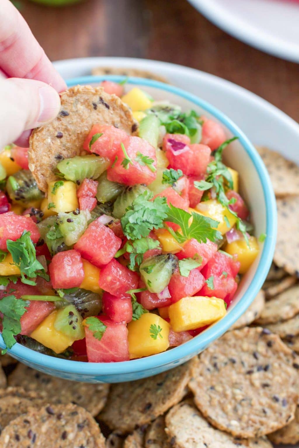A hand dipping a cracker into a bowl filled with watermelon mango salsa.
