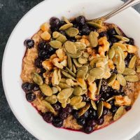 Vegan cinnamon oatmeal topped with nuts, seeds, and frozen blueberries in a white bowl.