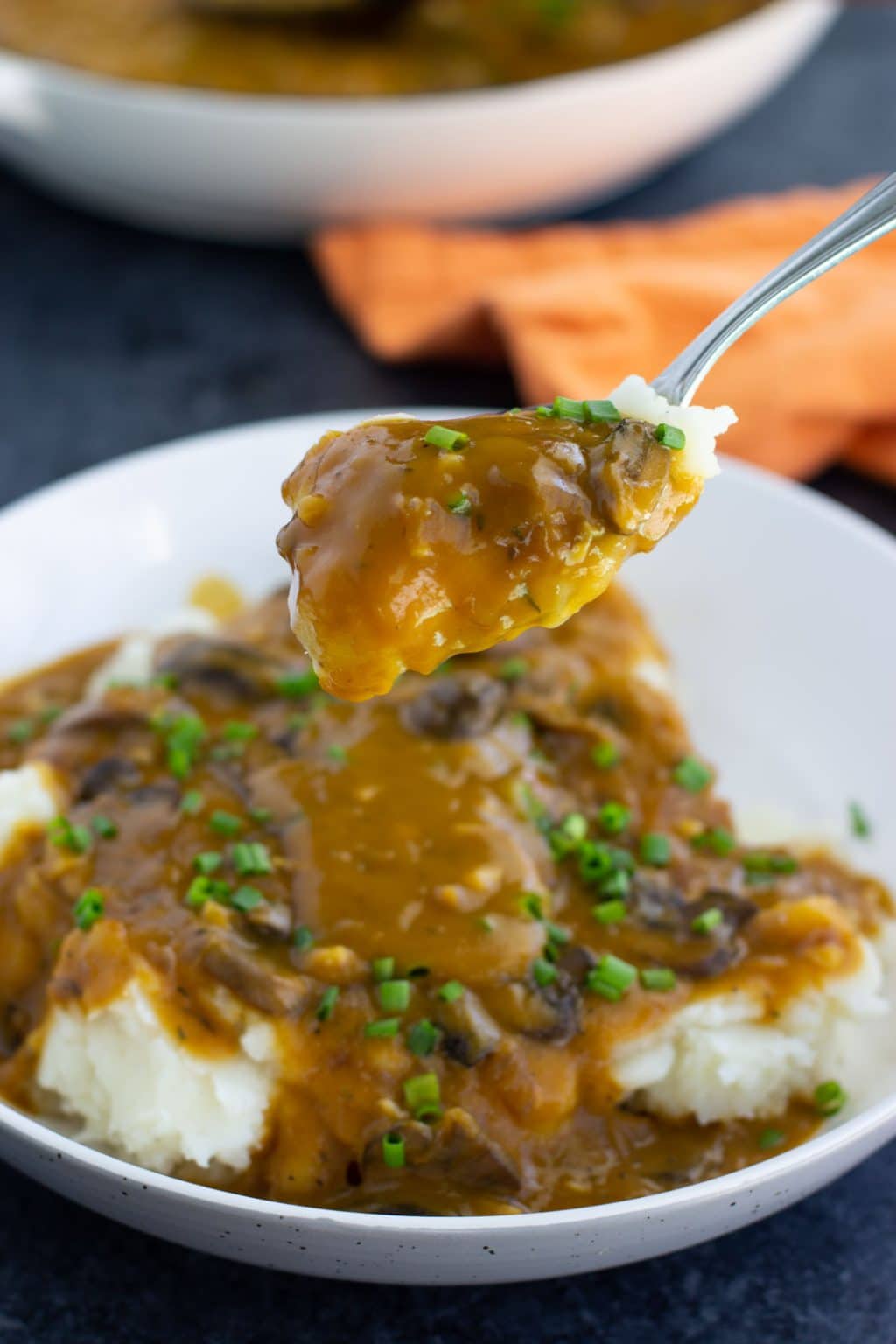 A spoonful of mashed potatoes topped with vegan gravy and chives over a white bowl next to an orange napkin on a dark background.