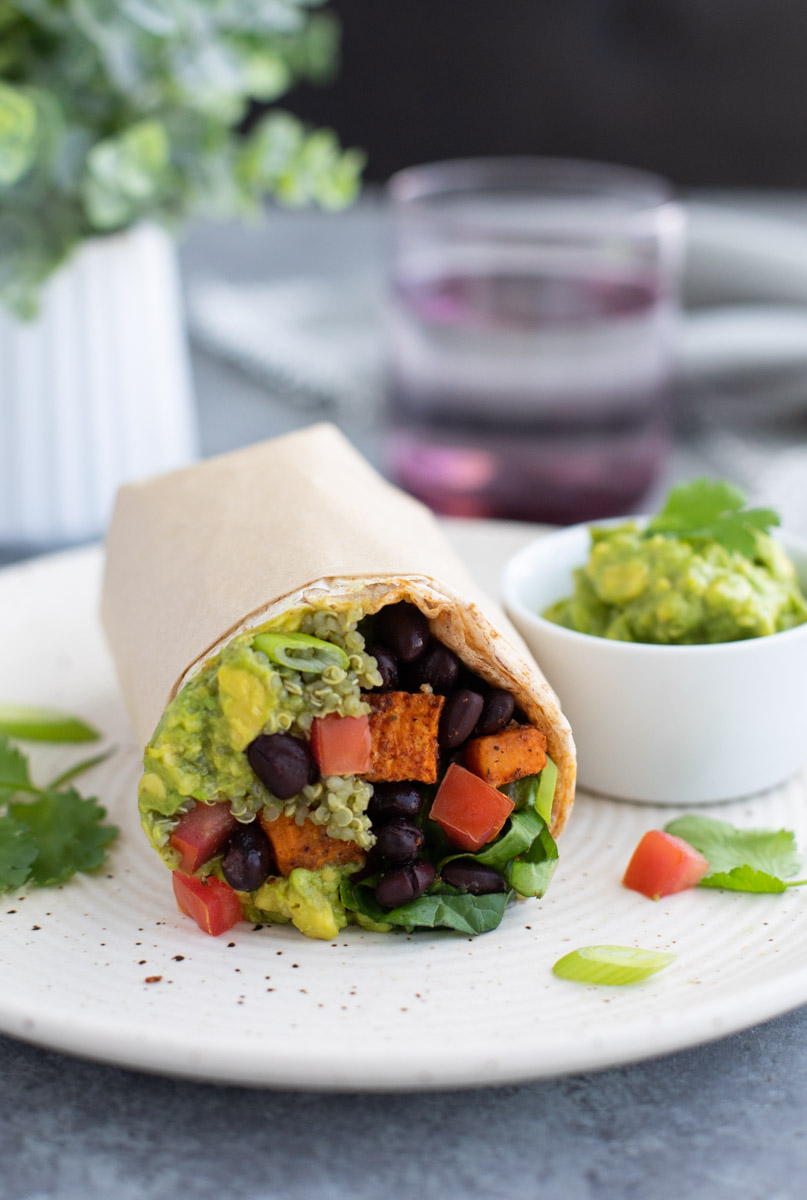 A close up view of a vegan burrito with a side of guacamole on a white plate.