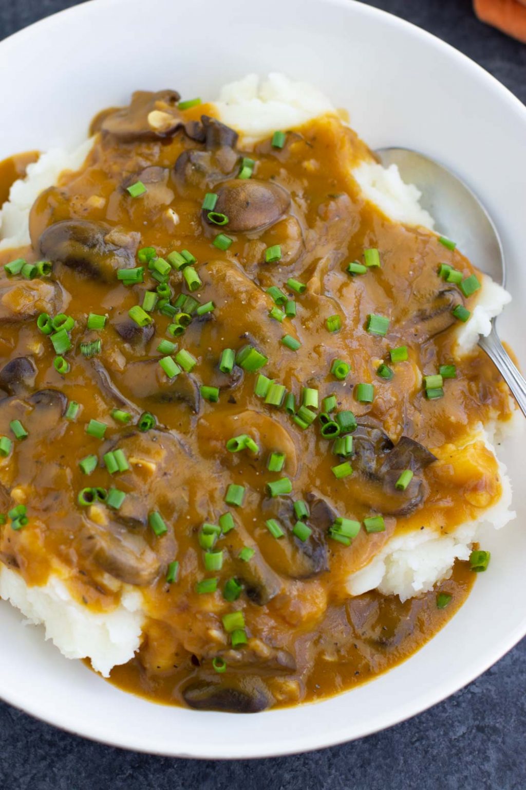 A white bowl filled with mashed potatoes, vegan mushroom gravy, chives, and a spoon on a dark background.