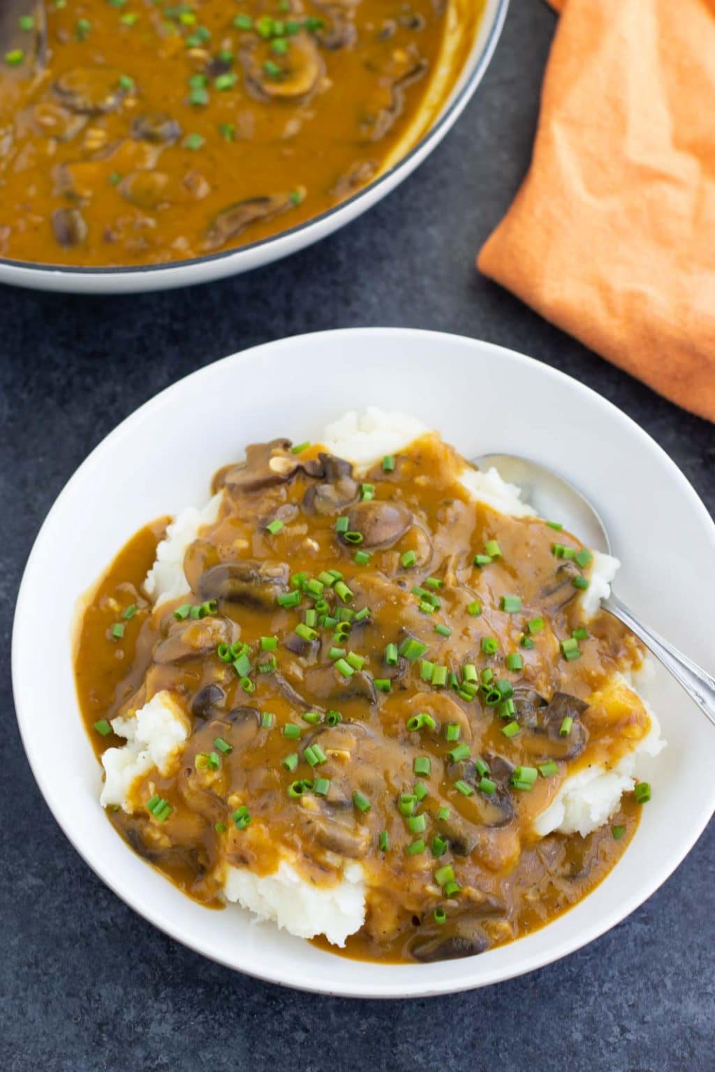 A white bowl filled with mashed potatoes, mushrooms, gravy, chives, and a spoon next to an orange napkin and a pan on a dark background.