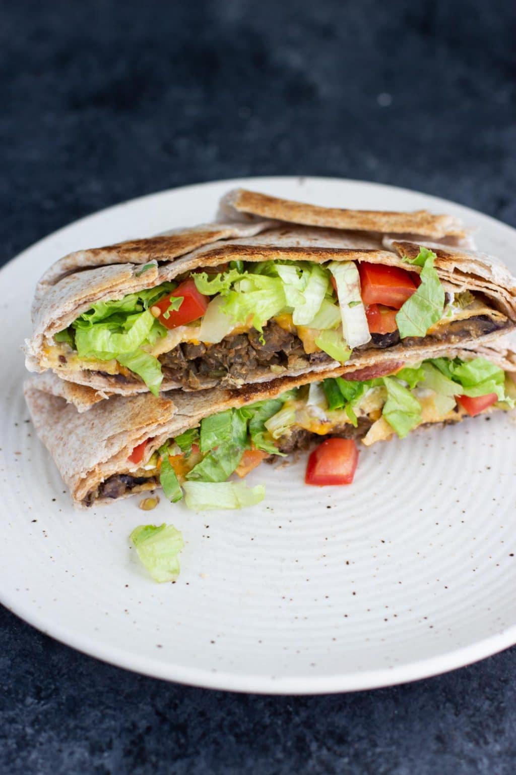 A vegan crunchwrap cut in half and stacked on top of each other on a white plate on a dark background. 