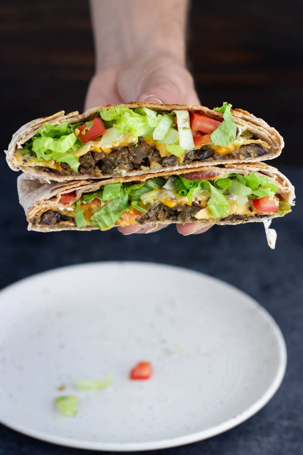 A hand holding two halves of a vegan crunchwrap supreme on top of each other over a white plate on a dark background.