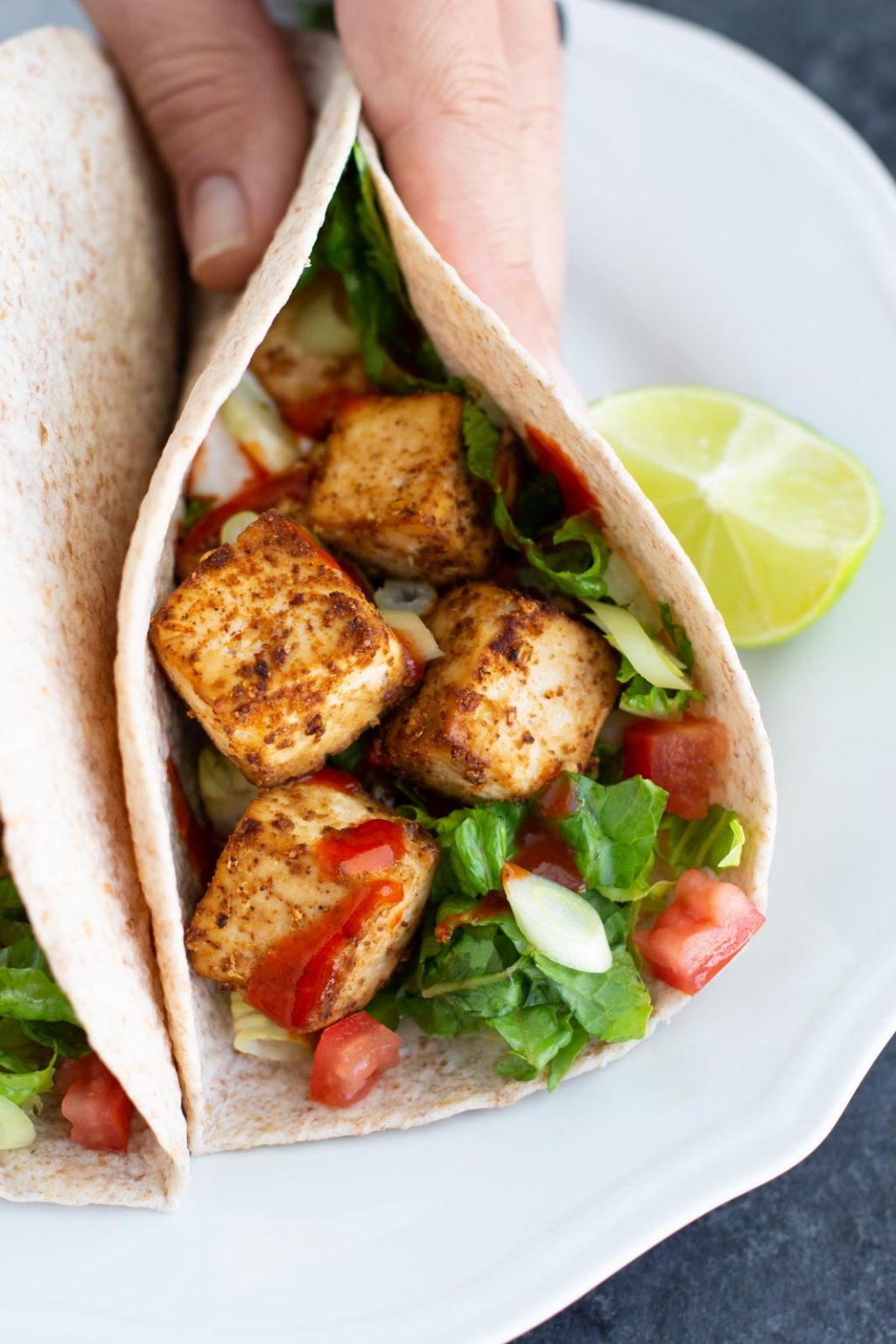 A hand holding a tofu taco on a white plate with a lime wedge. 