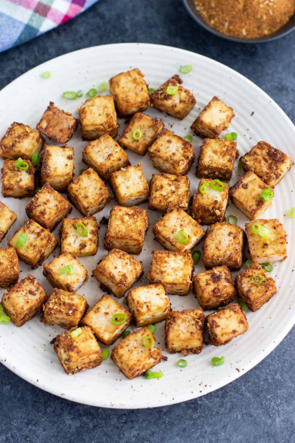 A white plate filled with tofu cubes next to a plaid napkin on a dark background. 