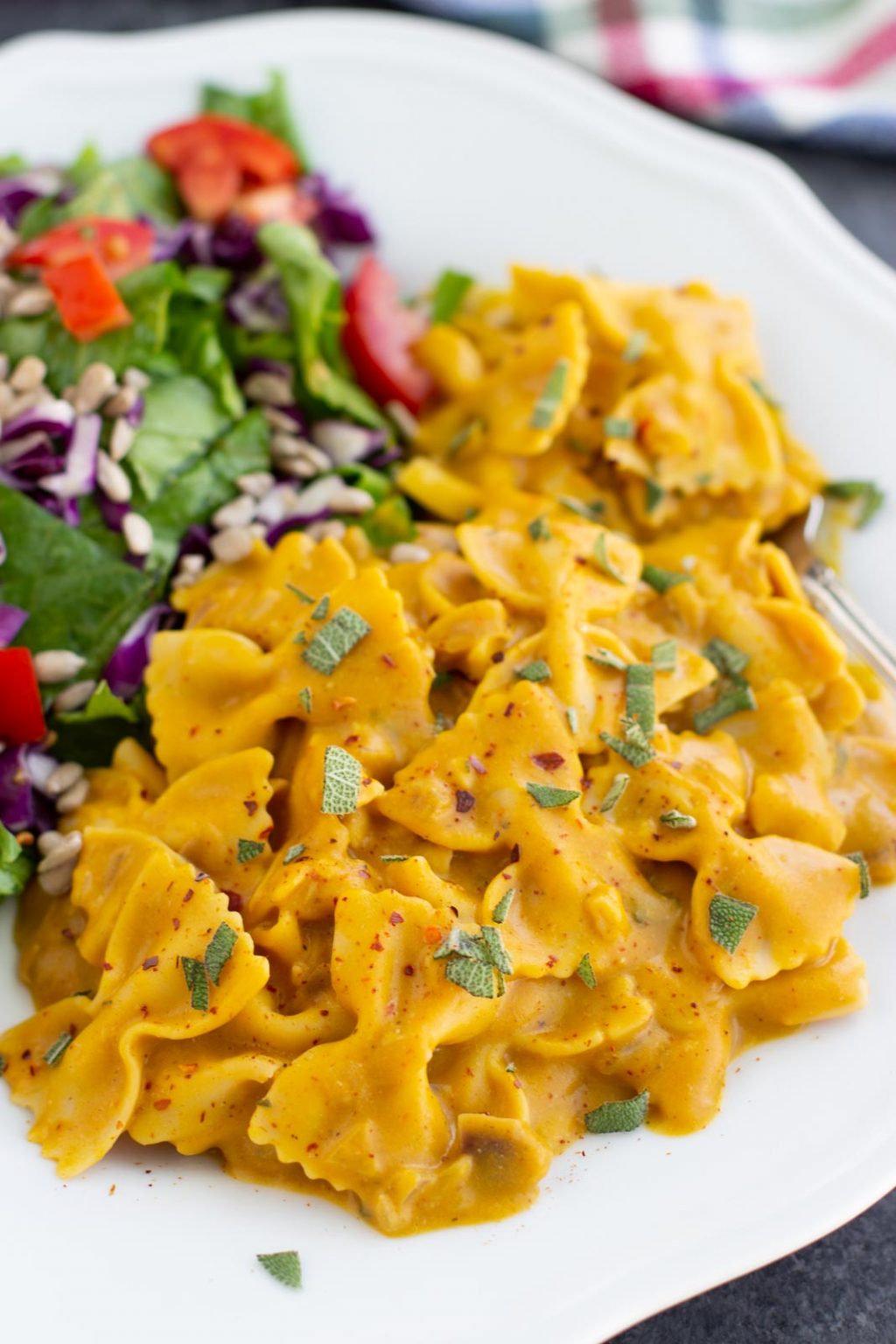 A white plate filled with pumpkin pasta and a romaine lettuce salad next to a plaid napkin.