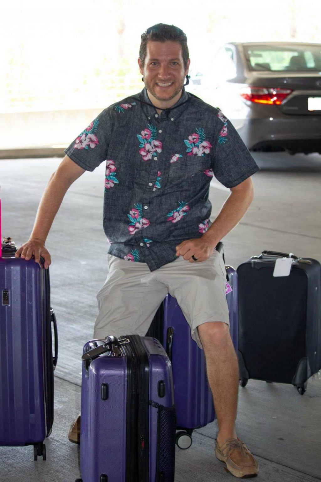 A man in a floral shirt is sitting on top of a purple suitcase with a few suitcases next to him.