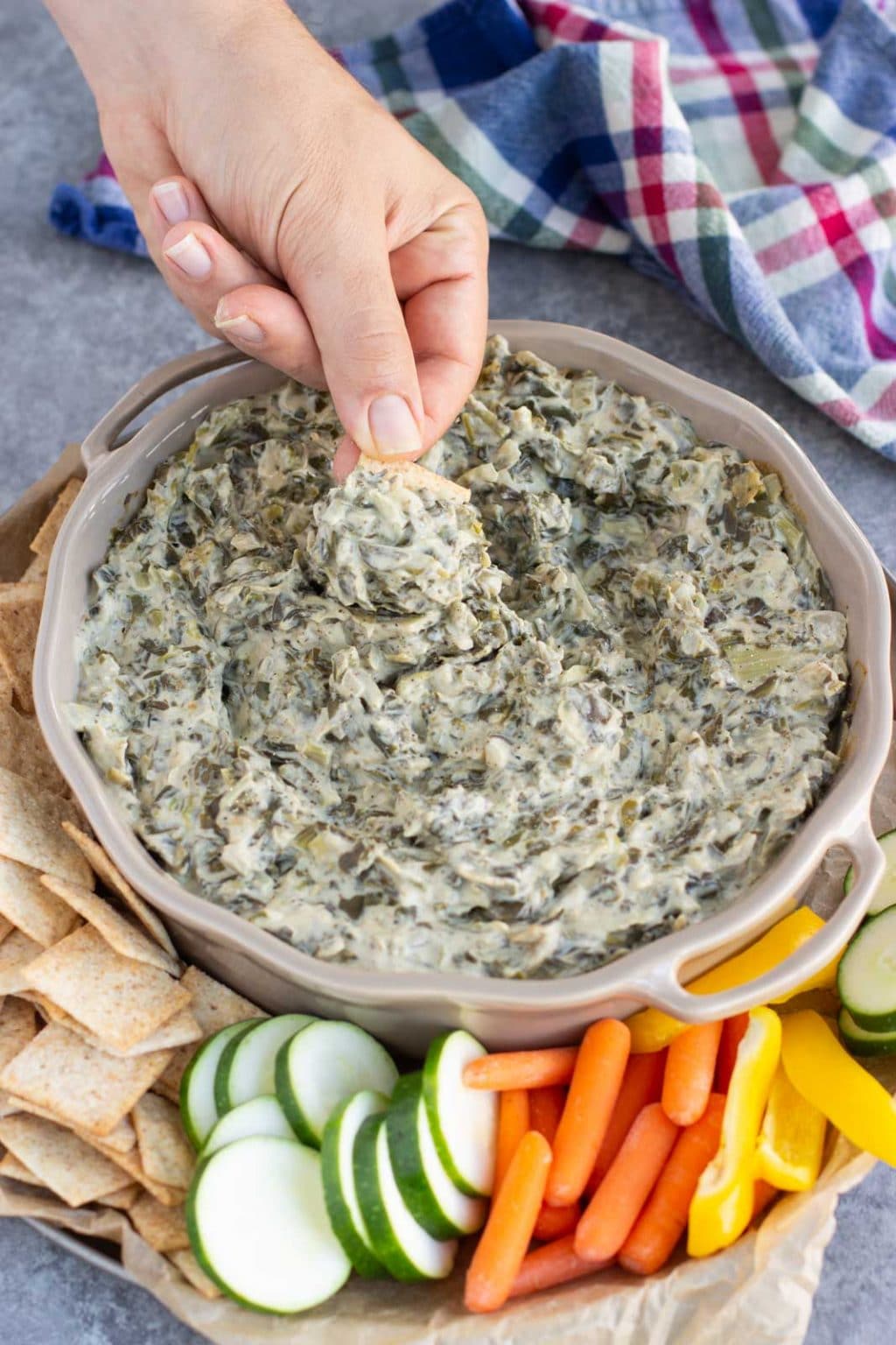 A hand dipping a cracker into a dish filled with spinach and artichoke dip next to a blue plaid towel.