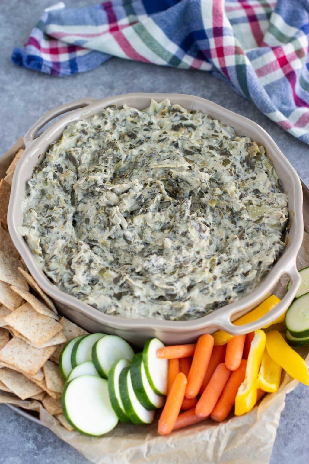 A cream colored dish filled with dip on a tray with veggies and crackers next to a blue plaid towel.