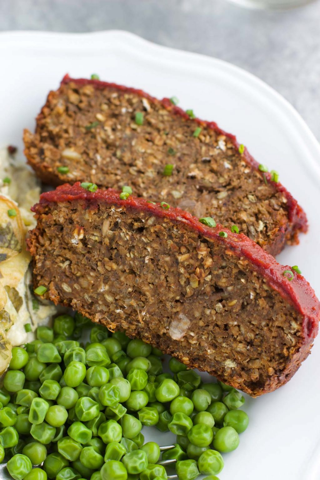 Two slices of vegan meatloaf next to potatoes and green peas on a white plate.