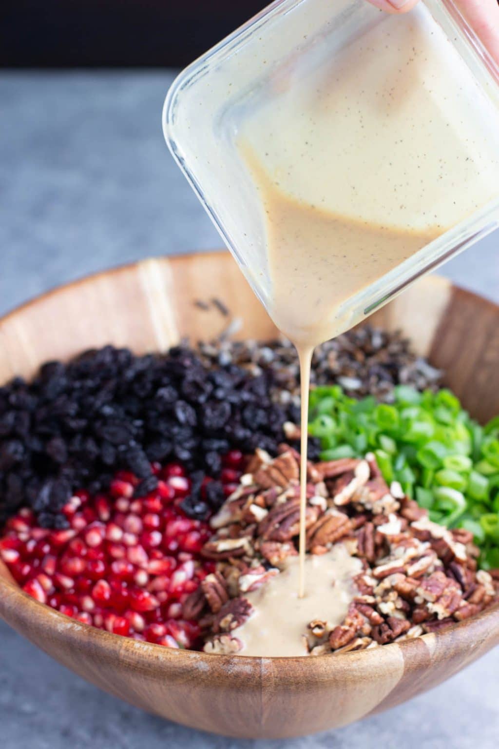 A hand pouring dressing over a wooden bowl filled with pomegranate arils, pecans, green onions, and dried cherries.