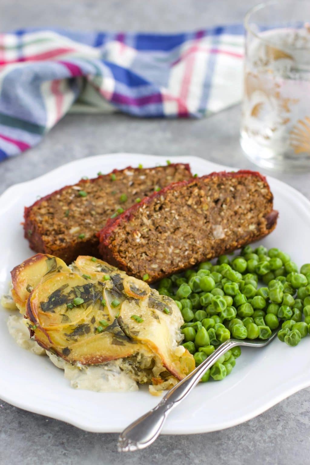 A white plate filled with scalloped potatoes, lentil loaf, and green peas next to a blue plaid napkin.