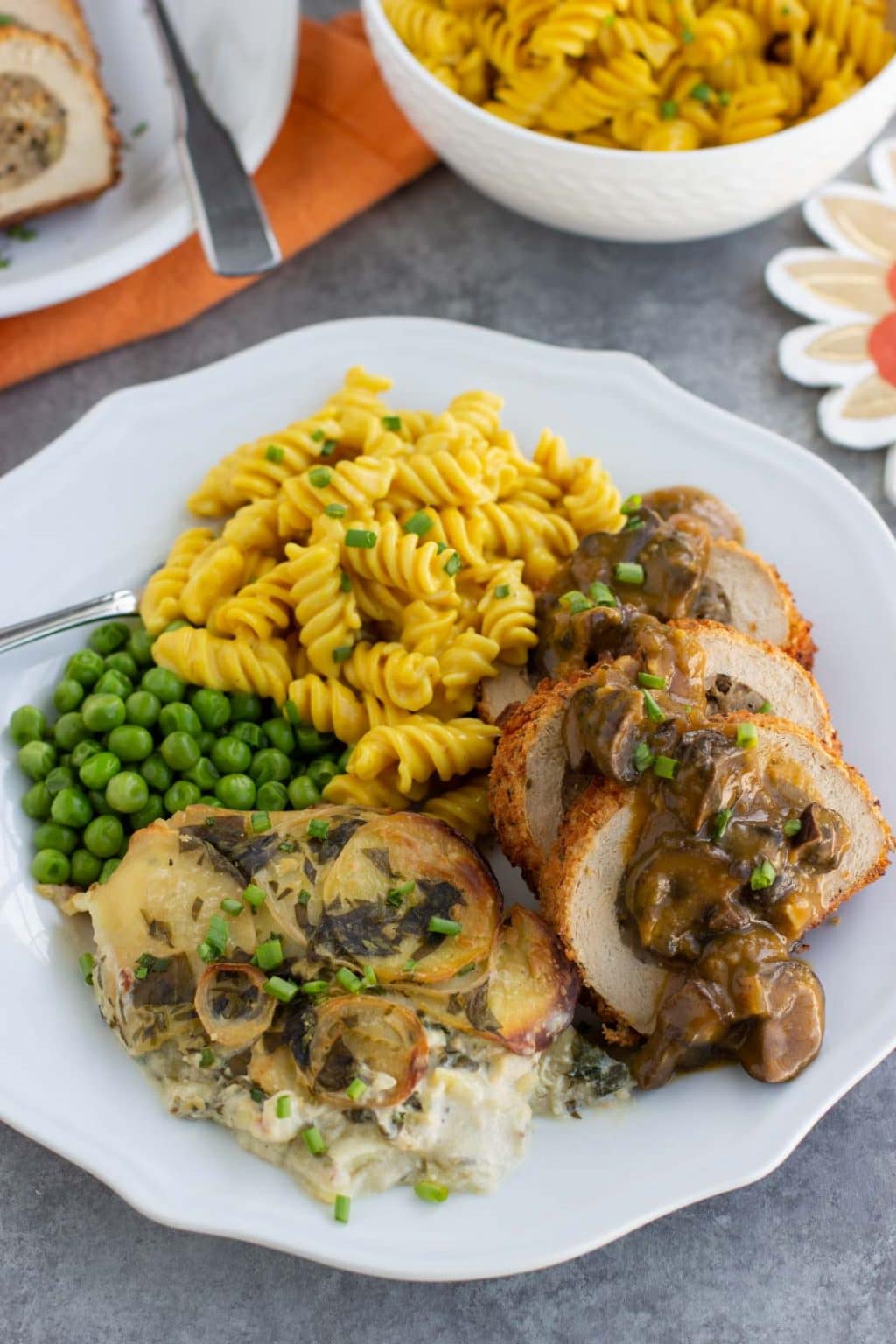 A white plate filled with scalloped potatoes, macaroni and cheese, vegan turkey, and green peas on a gray background. 