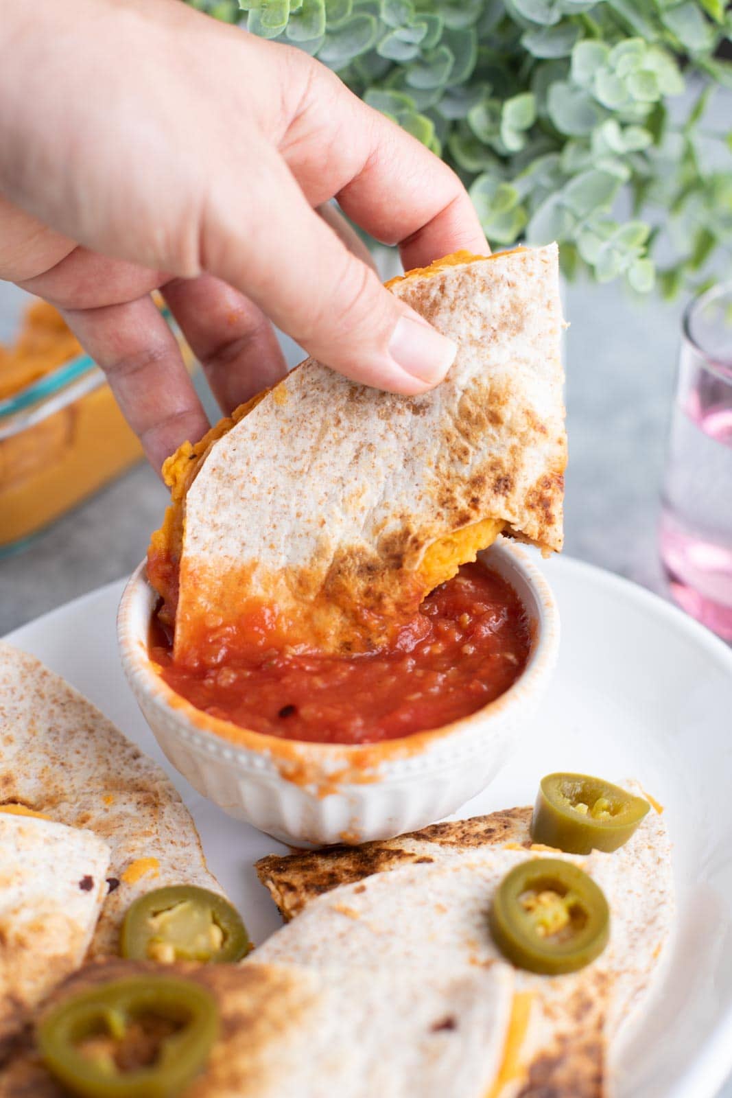 A hand dipping a quesadilla into a bowl of salsa on a white plate.