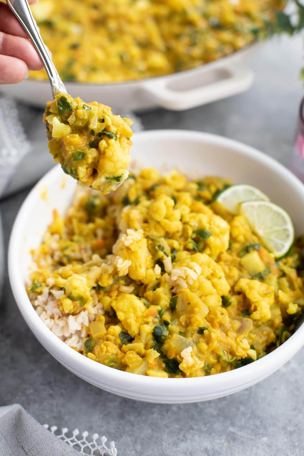 A hand holding a spoonful of lentil cauliflower curry over a bowl of brown rice. 