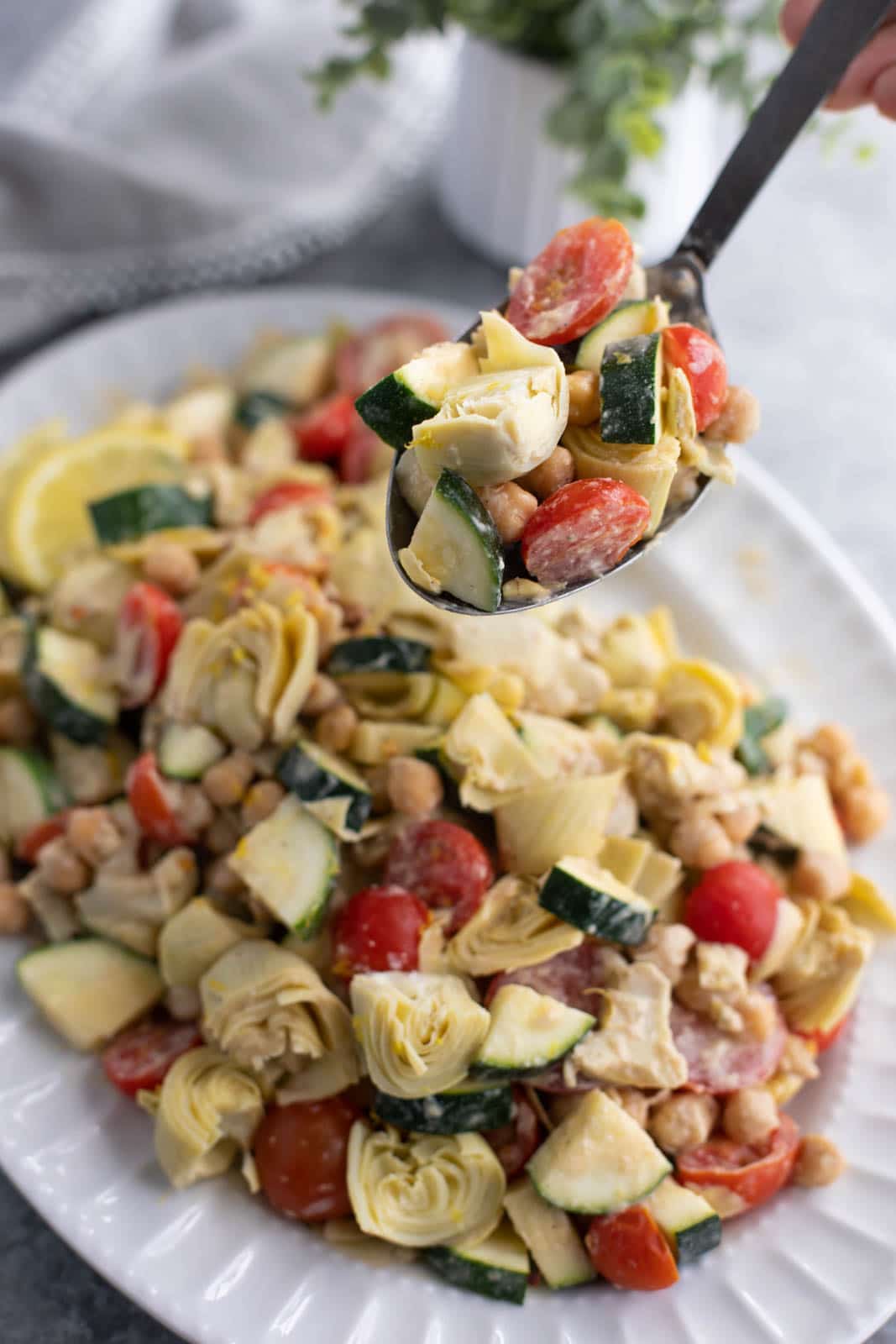 A hand holding a spoonful of zucchini salad over a large white platter.