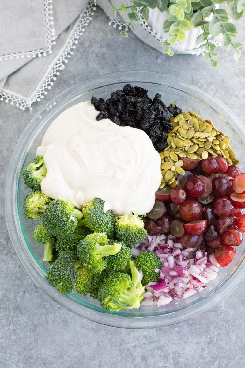 A large clear mixing bowl filled with broccoli, grapes, onions, pumpkin seeds, dried cherries, and creamy dressing on a gray background.