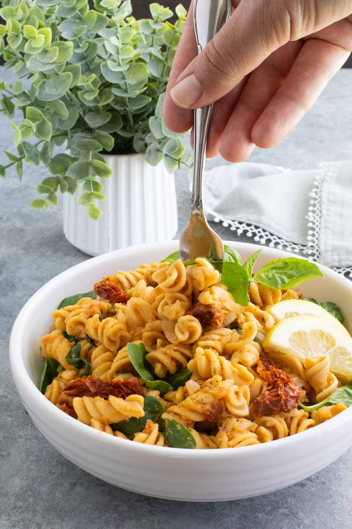 A hand pushing a fork into a white bowl filled with pasta in front of a small plant. 