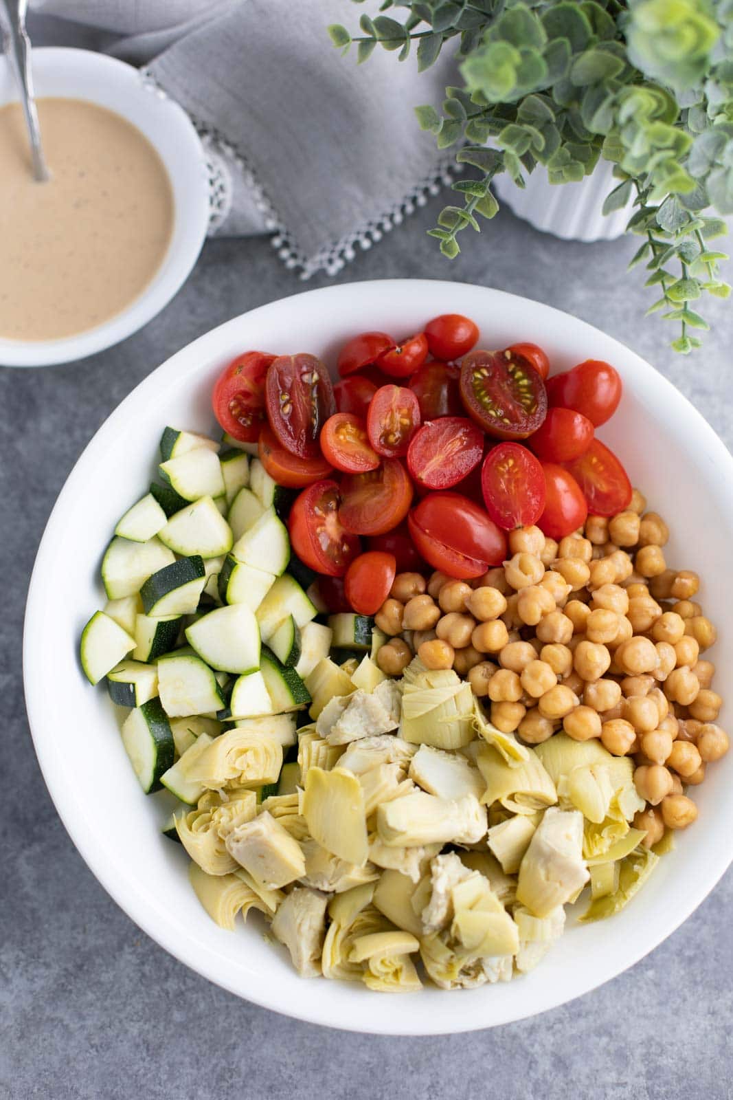 Salad ingredients in a large white bowl next to a plant and a bowl of lemon tahini sauce.